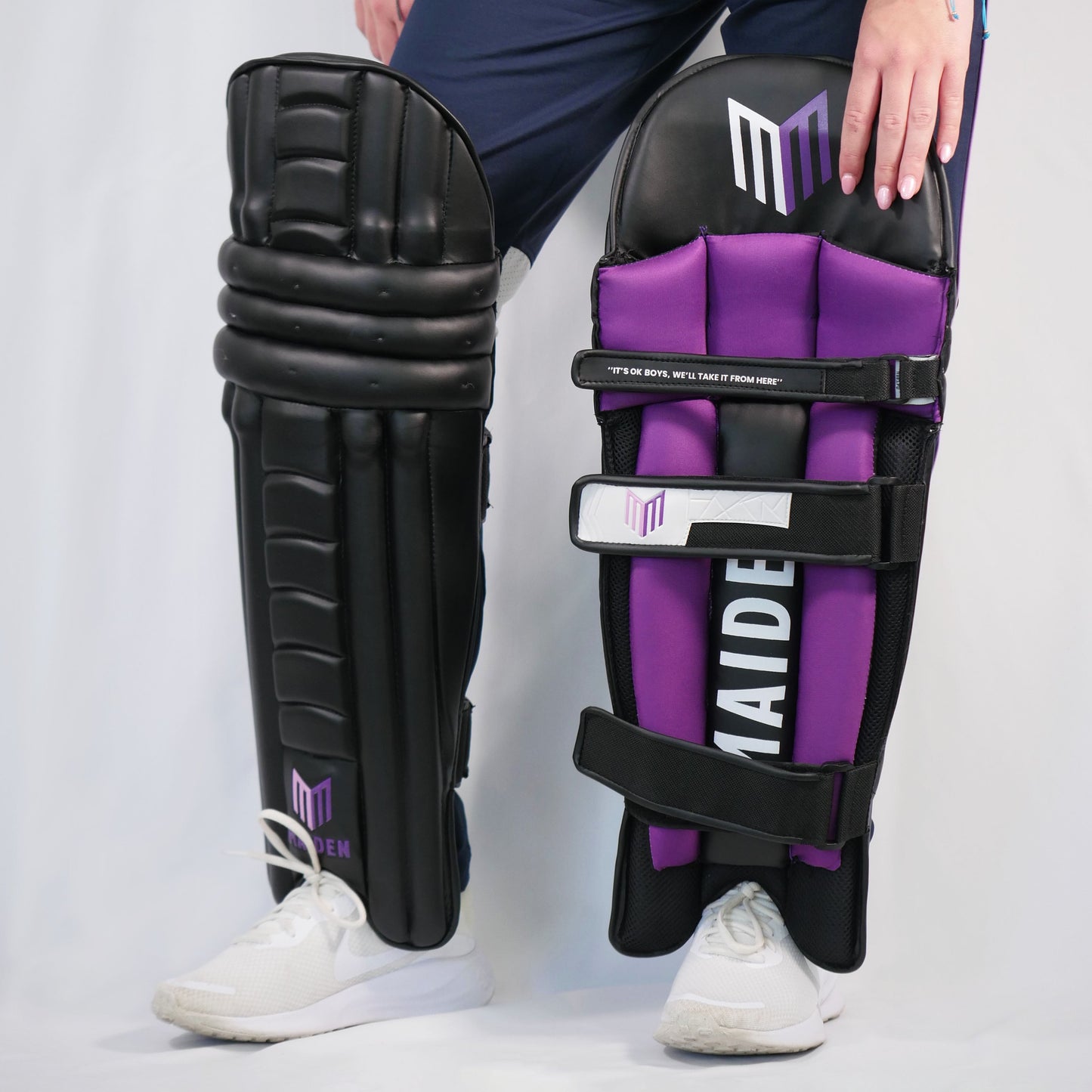 Person holding black and purple cricket leg guards with a visible brand logo on a white background
