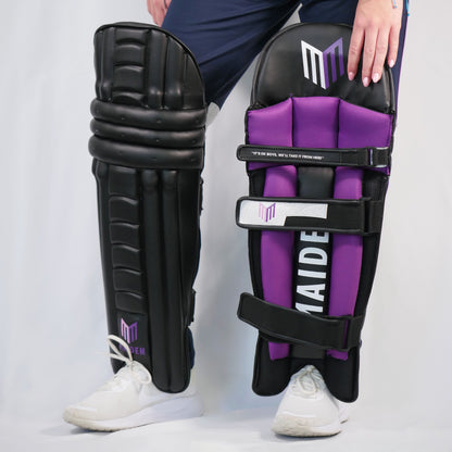 Person holding black and purple cricket leg guards with a visible brand logo on a white background
